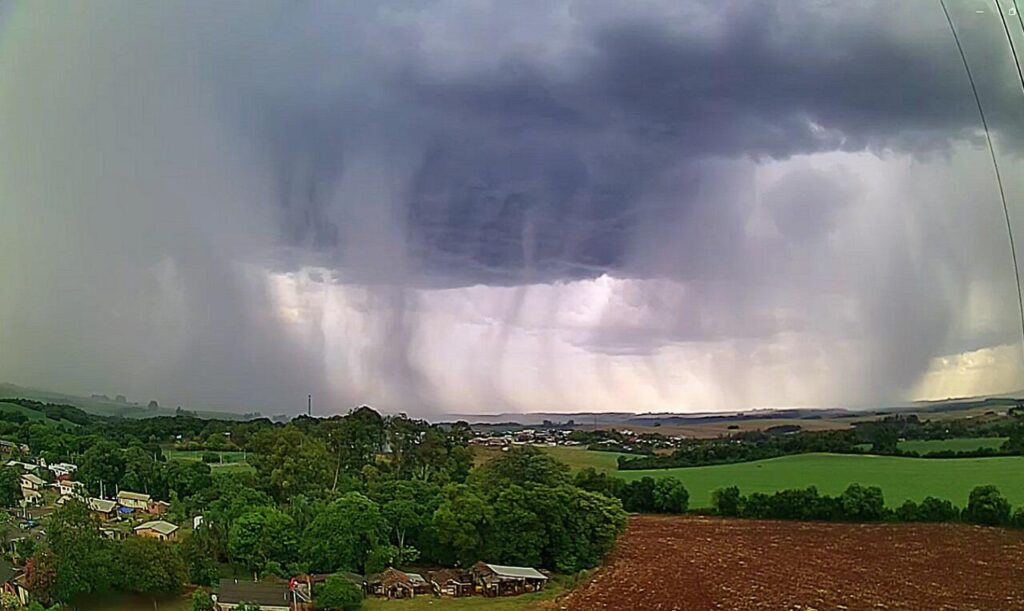 A foto mostra árvores e lavouras e o céu com nuvens e chuva forte